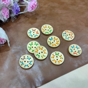 May include: Ten round wooden buttons with colourful embroidery. The buttons are beige with a light brown centre and have a cross-stitch pattern in shades of blue, green, orange, and yellow.
