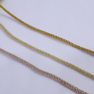 May include: Three metallic braided cords in gold, light gold, and rose gold are displayed against a white backdrop. The cords are arranged diagonally. The braided texture is visible, and the cords appear to be part of a craft or jewellery supply.