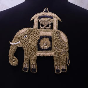 Könnte beinhalten: Gestickter Elefantenaufnäher in Gold und Marineblau. Der Elefant hat einen dekorativen Hodscha auf dem Rücken. Der Aufnäher ist mit aufwendiger Perlenarbeit und Metallfaden gefertigt und auf einer schwarzen Oberfläche befestigt.