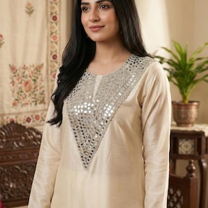 Puede incluir: Túnica crema de manga larga con escote en V adornado con detalles de espejo. La túnica está hecha de una tela suave y tiene un ajuste relajado. La modelo lleva la túnica. El fondo incluye un tapiz floral y una planta en maceta.