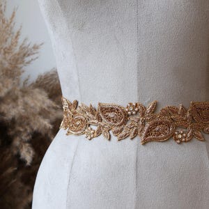 以下が含まれることがあります： 金色の刺繍が施されたベルトで、花とペイズリー柄の複雑な金属糸の細工が特徴です。小さな白いビーズと透明なクリスタルで装飾されており、明るいグレーの布で覆われたトルソーのマネキンに巻き付けられています。