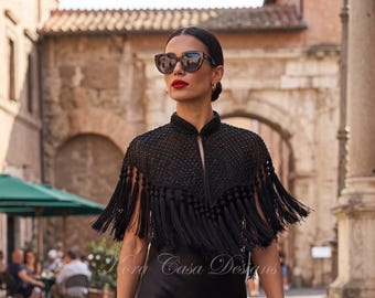 May include: A black capelet featuring a high mandarin collar, intricate beaded detailing across the shoulders, and long, flowing fringe trim along the hemline. The garment is adorned with the text Kora Casa Designs in a script font.