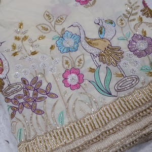 Peut inclure: Tissu ivoire avec broderies complexes représentant des oiseaux, des fleurs et du feuillage en rose, bleu, or et violet. Le motif comprend des bordures perlées et des ornements détaillés, créant un textile luxueux.