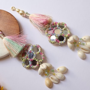 May include: Two decorative tassels with a floral design, mirrors, and seashells. Each tassel features a beaded top, a colorful tassel, and a flower-shaped embellishment with small mirrors and colorful beads. White seashells dangle from the bottom.
