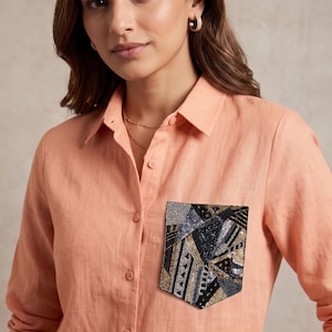 May include: A peach-colored long-sleeved button-down shirt featuring a single chest pocket adorned with a geometric pattern of black, silver, and gold beads and sequins. The shirt has a classic collar and a front button placket with matching buttons.
