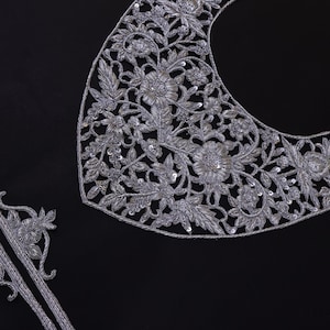 Può includere: Applicazione in pizzo ricamato argentato con motivi floreali e a foglie. L'applicazione è in due pezzi, un pezzo curvo per lo scollo e un pezzo di rifinitura dritto, entrambi con dettagli intricati. Lo sfondo è nero.