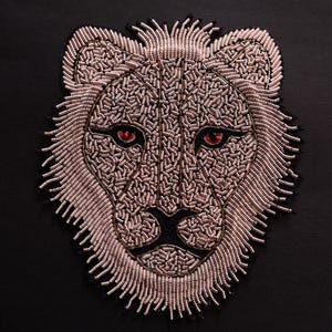 May include: A beaded lion's head with a pink and gold color scheme. The lion's eyes are red and the background is black.
