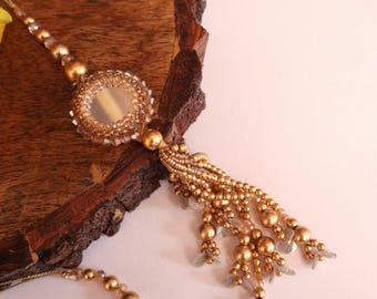 May include: A gold beaded necklace with a round mirror charm and a tassel. The necklace is on a wooden surface.