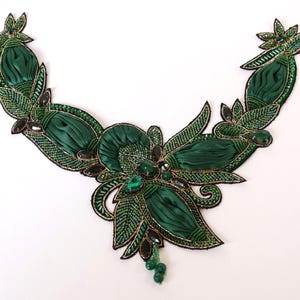 May include: A decorative collar appliqué in deep emerald green, featuring pleated satin fabric sections, intricate beadwork, and dark green faceted crystals. The design includes leaf-shaped beaded motifs and a small dangling beaded element at the centre.