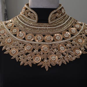 May include: A gold and white beaded and sequined collar necklace with a floral design. The necklace is on a black background.