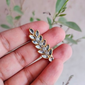 May include: A gold-toned leaf-shaped embellishment with clear, oval-shaped crystals and small, round, clear crystals. The crystals are set in a gold metal frame. The embellishment is held in a hand against a blurred green and beige background.