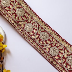 Op de afbeelding: Een decoratief bordeauxrood lint met gouden bloemenborduurwerk. Het lint heeft een herhalend patroon van bloemen en bladeren, met een rand van gouden versieringen. Het lint staat schuin in het frame, met gele bloemen in de linkerbenedenhoek.