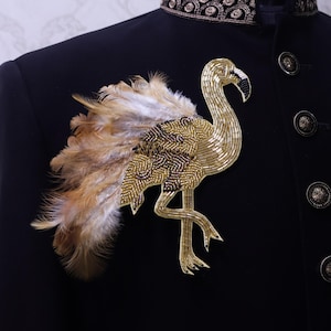 May include: A gold beaded flamingo brooch with feather accents, attached to a black jacket. The brooch features intricate beadwork and a detailed design. The feathers are a mix of brown and white. The jacket has decorative buttons.
