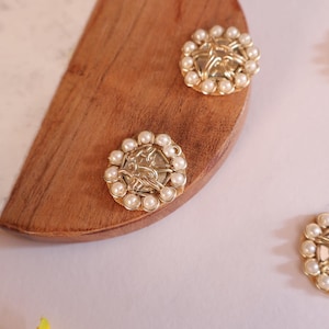 May include: Four gold-toned round embellishments with a pearl-like design. Each embellishment has a raised, intricate pattern in the centre. The embellishments are on a wooden surface.
