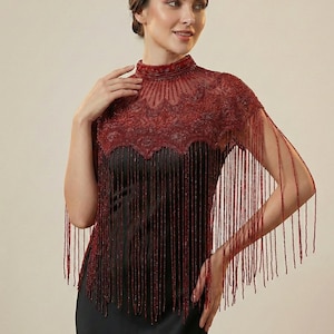 Op de afbeelding: Een bordeauxrode, met kralen bezette cape met een hoge kraag en franjedetails. De cape is versierd met ingewikkeld kralenwerk en hangt over een zwarte jurk. Het ontwerp heeft een geschulpte rand en lange kralenfranjes.