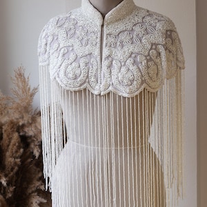 Op de afbeelding: Een wit kraaltjes-capelet met een hoge kraag en ingewikkeld bloemenborduurwerk. Het kledingstuk is versierd met dichte, lange witte kralenfranjes die vanaf de geschulpte zoom naar beneden hangen en het bovenlichaam bedekken met kralen.