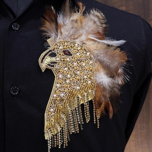 May include: A gold and brown eagle-shaped brooch with feathers and rhinestones. The brooch is attached to a black shirt.
