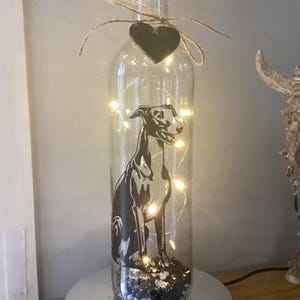 May include: A decorative glass bottle with a cork, featuring a black dog silhouette and fairy lights. A small black heart-shaped tag is tied to the bottle's neck. The bottle is displayed on a silver-coloured rotating base.
