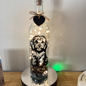 May include: A clear glass bottle with a cork stopper and a black heart charm. Inside the bottle is a black and white silhouette of a dog, surrounded by fairy lights. The bottle is sitting on a black base with a mirrored surface.