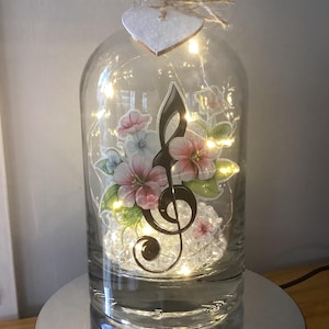 May include: A decorative glass bottle with a cork stopper, featuring a musical note and floral design. The bottle is lit by fairy lights, and a heart-shaped ornament hangs from the top. The bottle sits on a silver base. The bottle is likely around 25 cm tall.