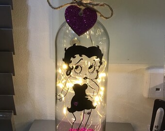 May include: A glass bottle with a pink glitter heart on the top and a black and white silhouette of a cartoon woman inside. The bottle is filled with pink glitter and white fairy lights.