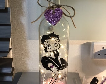 May include: A clear glass bottle with a cork top and a purple glitter heart tied to the neck. Inside the bottle is a black silhouette of a cartoon woman with the name "Helen" written below her. The bottle is filled with pink glitter and white fairy lights.