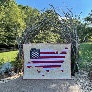 May include: A red, white and blue patchwork quilt in the shape of the United States of America. The quilt is displayed in front of a natural archway made of branches.