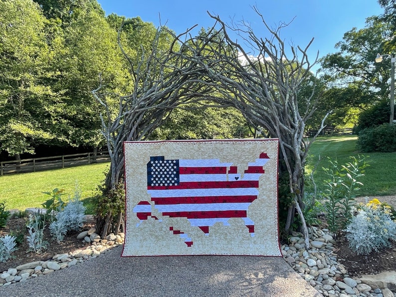 May include: A red, white and blue patchwork quilt in the shape of the United States of America. The quilt is displayed in front of a natural archway made of branches.