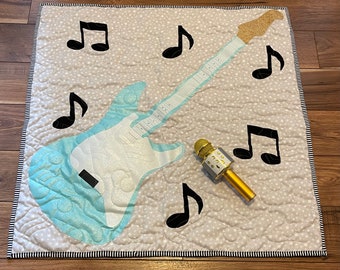 Könnte beinhalten: Eine hellblaue und weiße Gitarre mit goldenem Kopf und einem schwarzen Mikrofon auf einem grauen und weißen gesteppten Stoff mit schwarzen Noten.
