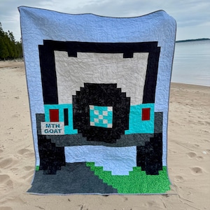 Può includere: Una trapunta pixelata con un design di veicolo fuoristrada blu e nero. La trapunta ha uno sfondo azzurro con un motivo ripetuto. Il veicolo ha accenti turchesi, uno pneumatico nero e il testo "MTN GOAT".