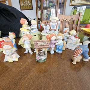 May include: A collection of vintage porcelain figurines, featuring children in various poses and outfits. The figurines are painted in pastel colors and include a wishing well with the text "Wishes 20¢". The collection is displayed on a wooden table.