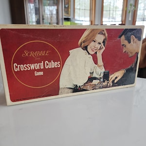 May include: Vintage Scrabble Crossword Cubes game box. The box has a red background with the game title in white and a circular logo. The box features an illustration of a smiling woman and a man playing the game.