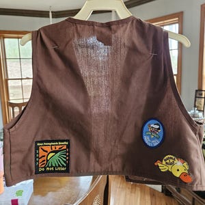 May include: A brown vest with three patches. One patch reads "Keep Pennsylvania Beautiful" with "Do Not Litter" below. Another patch says "Girl Scout!" and "Product Sale". A third patch features a yellow duck with the words "Waddle Head?"