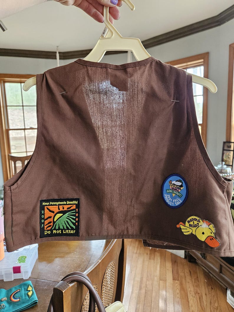 May include: A brown vest with three patches. One patch reads "Keep Pennsylvania Beautiful" with "Do Not Litter" below. Another patch says "Girl Scout!" and "Product Sale". A third patch features a yellow duck with the words "Waddle Head?"