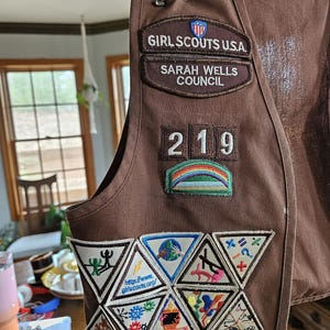 May include: A brown Girl Scouts vest with multiple patches. The vest has patches that read "GIRL SCOUTS U.S.A.", "SARAH WELLS COUNCIL", and "219". Several triangular patches with various designs and the website "http://www.girlscouts.org/" are also visible.