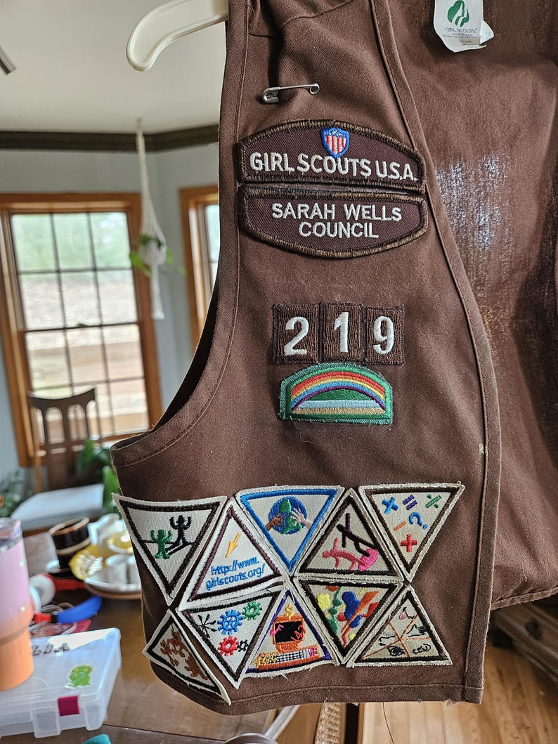 May include: A brown Girl Scouts vest with multiple patches. The vest has patches that read "GIRL SCOUTS U.S.A.", "SARAH WELLS COUNCIL", and "219". Several triangular patches with various designs and the website "http://www.girlscouts.org/" are also visible.