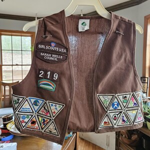 May include: Brown Girl Scouts USA vest with multiple patches and the text "GIRL SCOUTS USA SARAH WELLS COUNCIL 219". The vest has several triangular patches with various colorful designs and a rainbow patch.