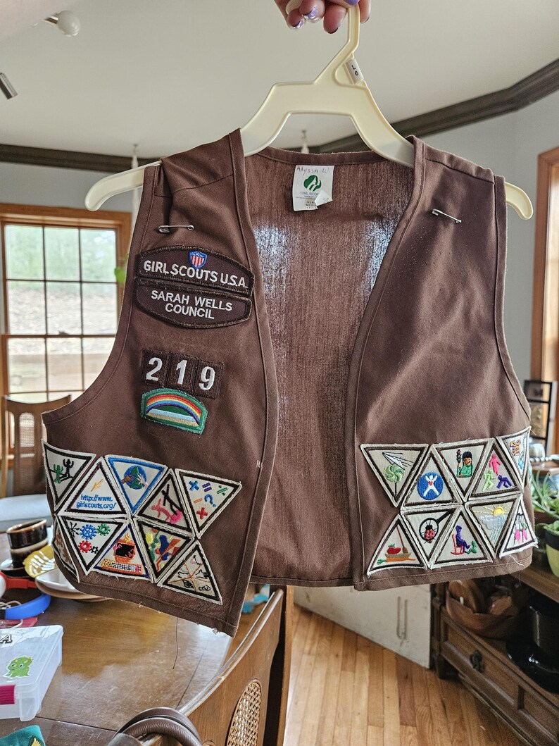 May include: Brown Girl Scouts USA vest with multiple patches and the text "GIRL SCOUTS USA SARAH WELLS COUNCIL 219". The vest has several triangular patches with various colorful designs and a rainbow patch.