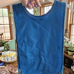 May include: A solid blue bib-style apron hanging on a white hanger. The apron has a rounded bottom and ties on the sides. The background includes a wooden table and a glimpse of a Girl Scout vest.