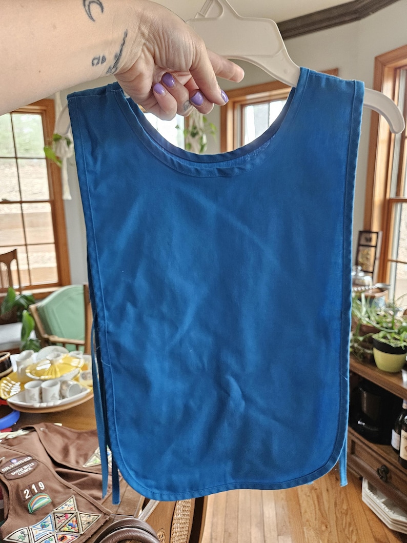 May include: A solid blue bib-style apron hanging on a white hanger. The apron has a rounded bottom and ties on the sides. The background includes a wooden table and a glimpse of a Girl Scout vest.