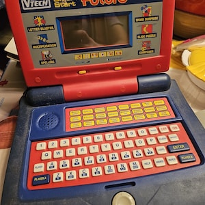 May include: A red and blue VTech Smart Start Future learning laptop. Features a keyboard, screen, and buttons for various educational games. Includes text like "Letter Blaster," "Multiplication," and "Word Surprise."