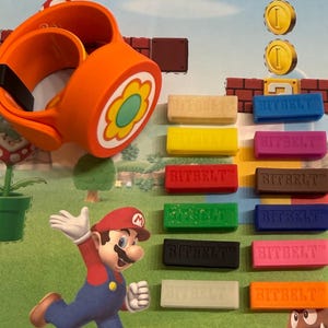 May include: An orange wearable device with a flower design, and a collection of colorful "BITBELT" bands. The image features a cartoon of Mario running, with coin graphics and a brick background, evoking a video game theme.