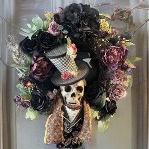 May include: A Halloween wreath with a skull wearing a top hat and a decorative jacket. The wreath is decorated with black and purple roses, dark green leaves, and other floral elements. The hat has a black and white checkered pattern.