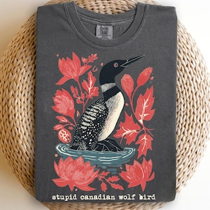 Pode incluir: T-shirt cinzenta escura Comfort Colors com um gráfico de um mergulhão na água, rodeado por flores e folhas vermelhas. O texto "stupid canadian wolf bird" está impresso na parte inferior. A camisa é exibida em uma superfície tecida.
