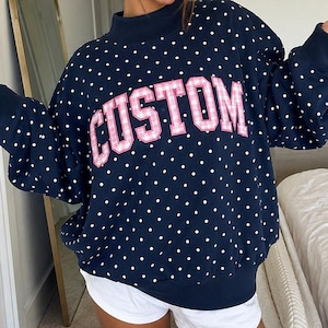 May include: A navy blue long-sleeve sweatshirt featuring a white polka dot pattern and a mock neck collar. The word CUSTOM is appliqued across the chest in pink and white gingham fabric with a white border, paired with plain white shorts.