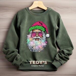 Op de afbeelding: Een olijfgroene sweatshirt met een afbeelding van de kerstman met een zonnebril en een roze glitterhoed. Zijn baard is versierd met kleurrijke lichtjes. De tekst "TEDY'S Creative Market" staat onderaan op de sweatshirt.