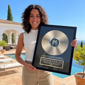 May include: A framed gold record award with a black border. The award features the text "ELEANOR STERLING" and "ASCENT DIGITAL HOLDINGS 2025". The background is a blue-green color.