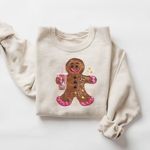 May include: A beige crewneck sweatshirt featuring a central graphic of a brown gingerbread person holding a pink cup with a straw. The gingerbread figure has pink icing accents on its hands and feet, black button eyes, and gold star details.