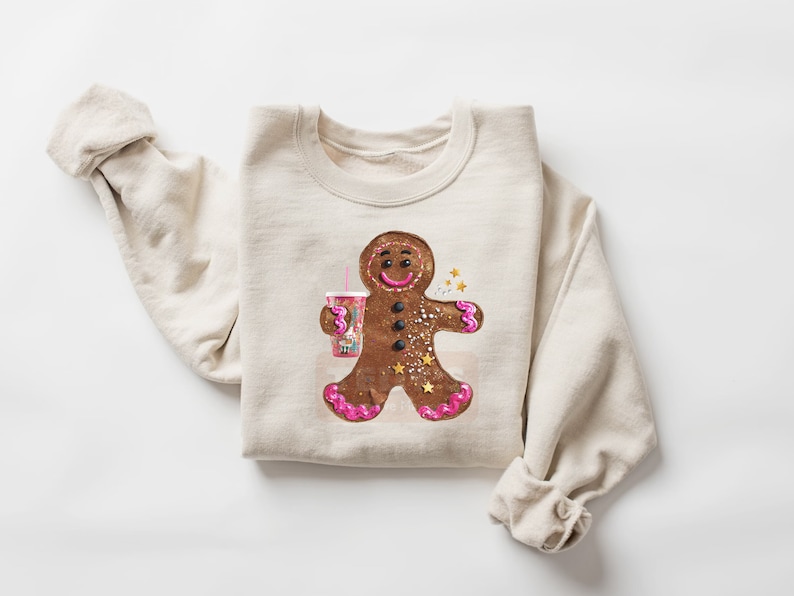 May include: A beige crewneck sweatshirt featuring a central graphic of a brown gingerbread person holding a pink cup with a straw. The gingerbread figure has pink icing accents on its hands and feet, black button eyes, and gold star details.