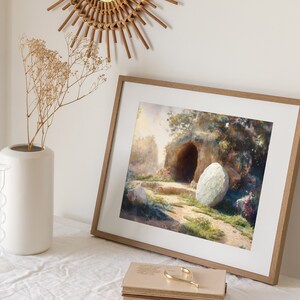 May include: A framed painting depicting a stone tomb with a large, round stone rolled away from the entrance. The tomb is surrounded by lush greenery and flowers, suggesting a peaceful and hopeful scene.